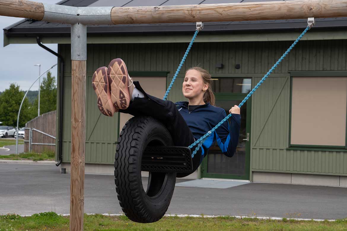 Nærbilde av en jente i tenårene som husker på en dekkhuske utendørs. Hun har på seg en mørkeblå treningsjakke og sorte bukser, og smiler mens hun svinger seg høyt opp med bena strakt ut foran seg. Husken henger i blå kjeder fra et stabilt trestativ. I bakgrunnen ser man en grønnmalt skolebygning med mørkt tak, og en asfaltert skolegård.
