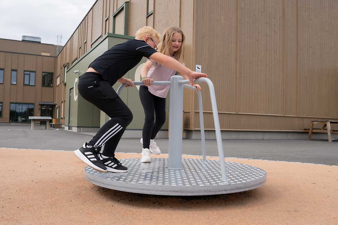 • En jente og en gutt i skolealder står oppå en rund, snurrende karusell av metall i en skolegård. Gutten har på seg sorte treningsklær med hvite striper og briller, og lener seg fremover mens han holder i metallbøylene. Jenta har på seg en lyserød t-skjorte og sorte tights, og holder også fast i stativet. Underlaget rundt karusellen er av lyst, mykt gummidekke, og i bakgrunnen ses en moderne skolebygning i tre.