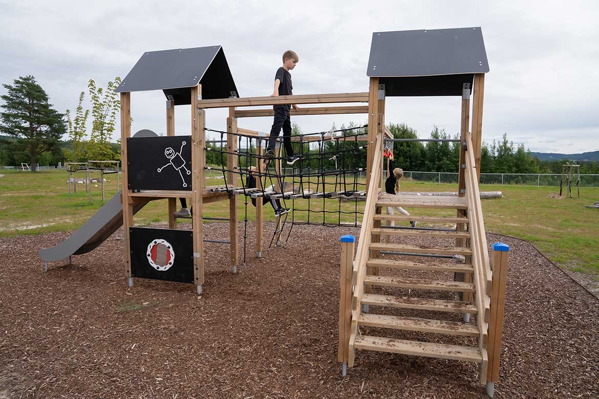 Gutt balanserer over en nettbro mellom to leketårn i tre med svarte tak. Lekestativet har trapp, sklie og klatrenett og står på en lekeplass utendørs.