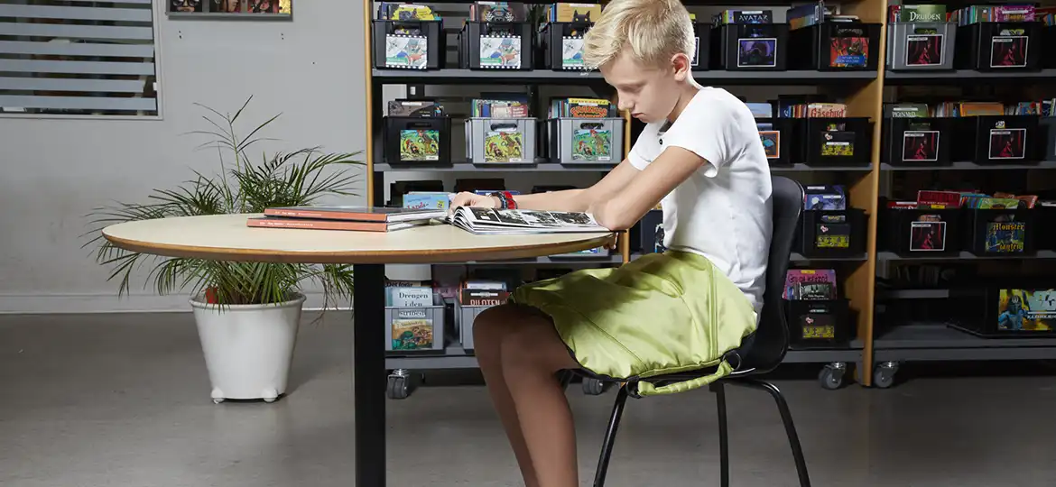 En gutt som sitter og leser på biblioteket og har en Protac KneedMe pute i fanget.