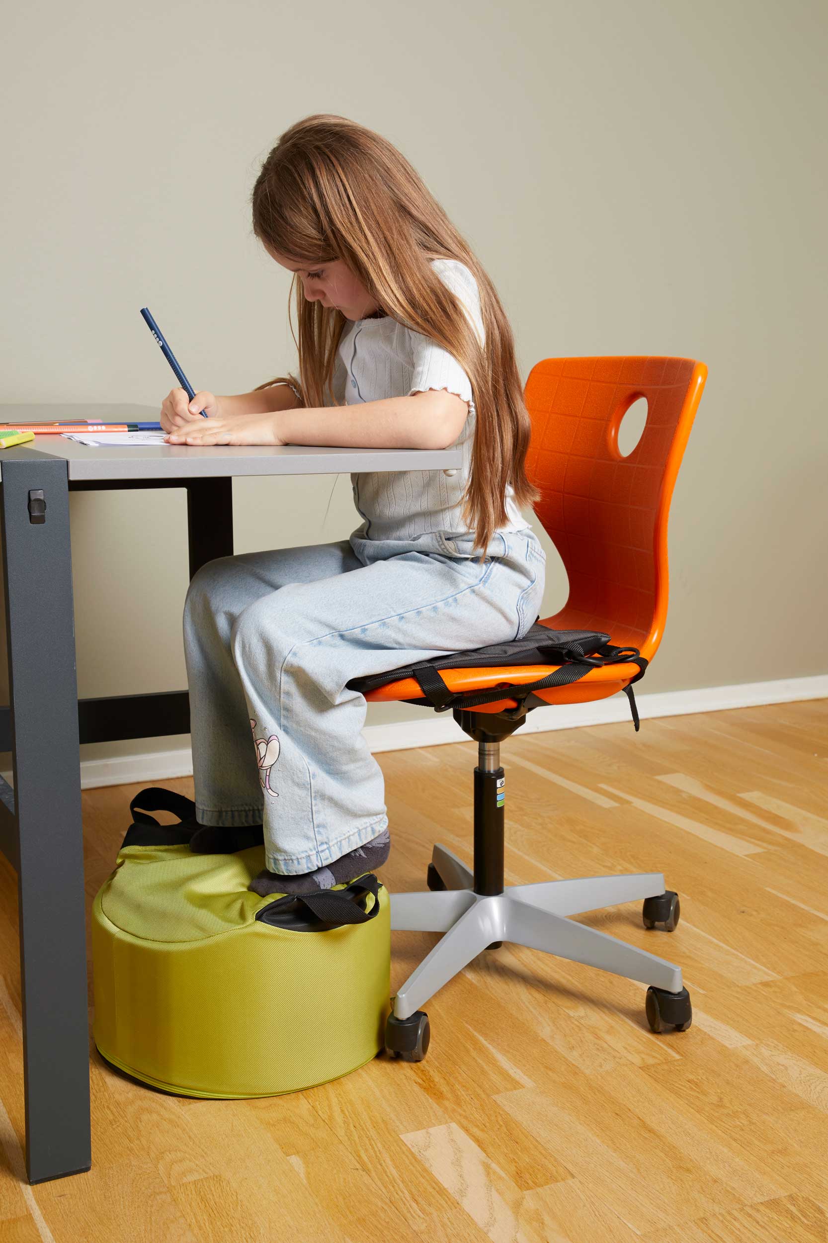 En elev sitter ved et skolebord og skriver. Eleven sitter på en oransje stol med en sittepute og har føttene plassert på en grønn fotstøtte.