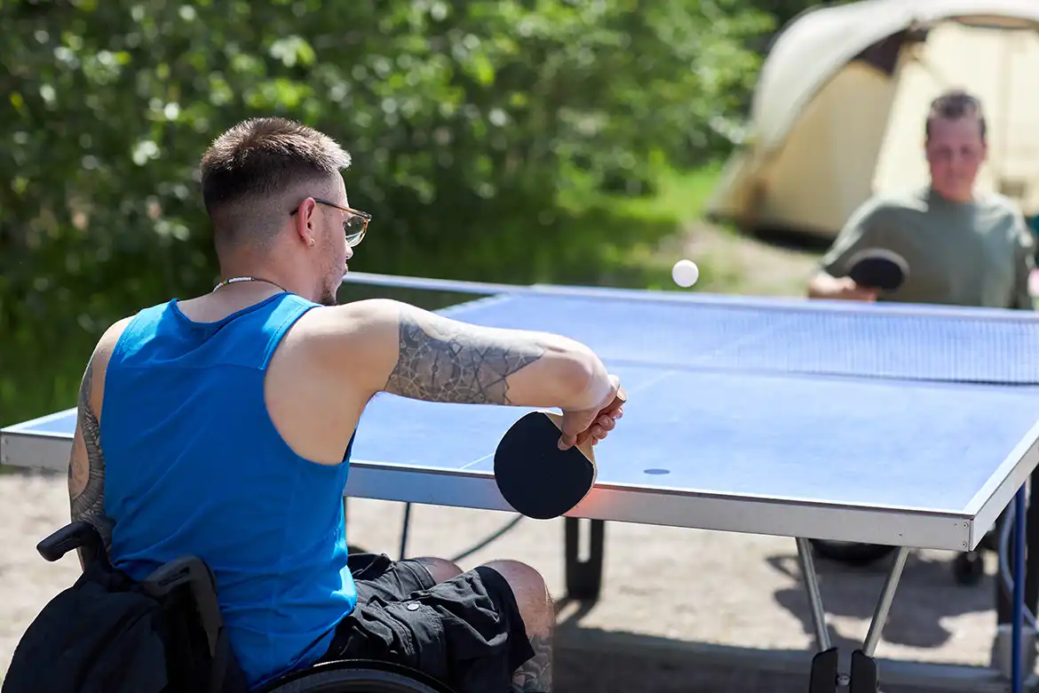 To gutter i rullestol spiller bordtennis utendørs i sola.